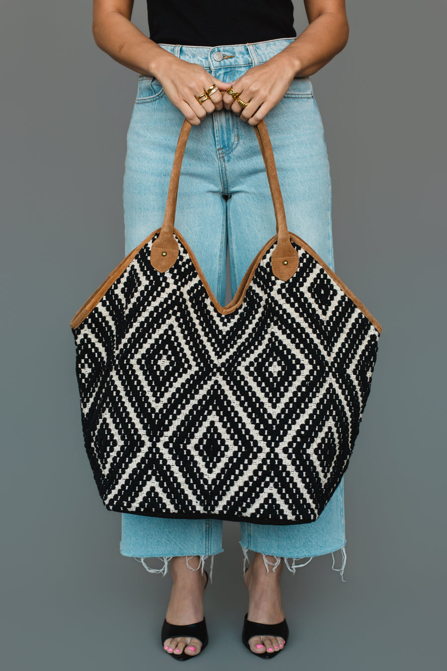 A person in light blue jeans holds the Panache Apparel Collection Black & Cream Diamond Pattern Textured Tote, featuring brown handles. Only their lower torso and legs are visible against a plain gray background.
