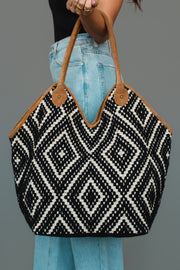 A person wearing light blue jeans and a black top holds the Panache Apparel Collection Black & Cream Diamond Pattern Textured Tote, which features black and white geometric diamond designs and brown handles, against a plain gray background.