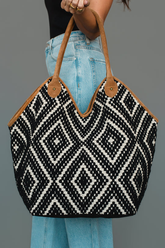 A person wearing light blue jeans and a black top holds the Panache Apparel Collection Black & Cream Diamond Pattern Textured Tote, which features black and white geometric diamond designs and brown handles, against a plain gray background.