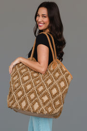 A woman with long dark hair, dressed in a black shirt and light blue jeans, smiles while carrying the Panache Apparel Collection Brown & Cream Diamond Pattern Textured Tote, featuring leather handles, as she stands against a plain gray background.