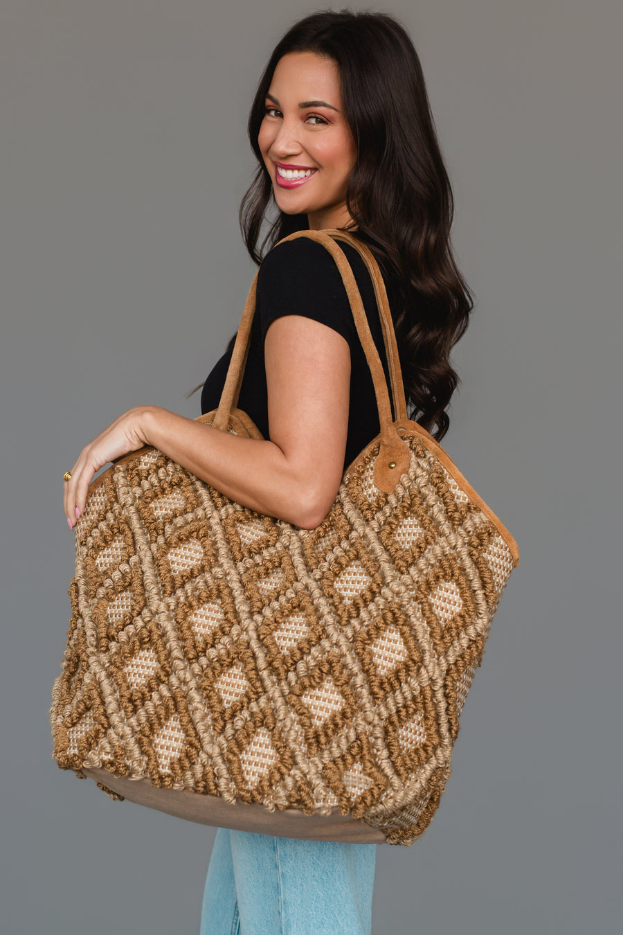 A woman with long dark hair, dressed in a black shirt and light blue jeans, smiles while carrying the Panache Apparel Collection Brown & Cream Diamond Pattern Textured Tote, featuring leather handles, as she stands against a plain gray background.