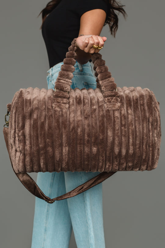 A person wearing a black shirt and blue jeans holds the Panache Apparel Collection Brown Super Soft Ribbed Duffel, featuring handles and a shoulder strap, against a gray background.