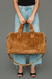 A person in a black top and light blue jeans holds the Mustard Super Soft Ribbed Duffel from Panache Apparel Collection with both hands. The duffel bag features two handles, a shoulder strap, and a ribbed faux fur texture. The person is wearing black open-toe heels.