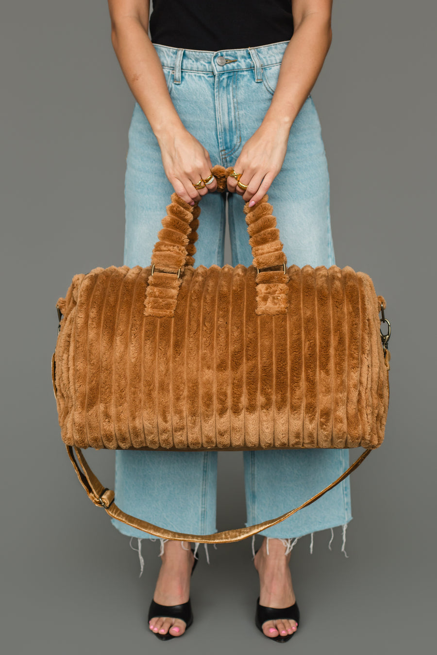 A person in a black top and light blue jeans holds the Mustard Super Soft Ribbed Duffel from Panache Apparel Collection with both hands. The duffel bag features two handles, a shoulder strap, and a ribbed faux fur texture. The person is wearing black open-toe heels.