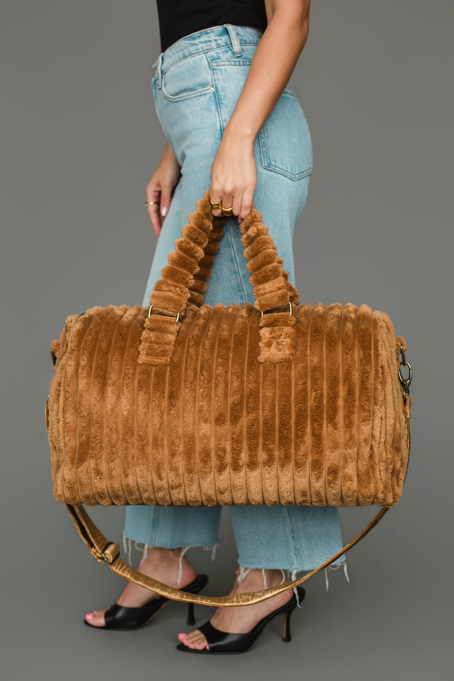 A person in blue jeans and black heels holds the Mustard Super Soft Ribbed Duffel from Panache Apparel Collection, featuring matching handles and a detachable shoulder strap, standing against a plain gray background.