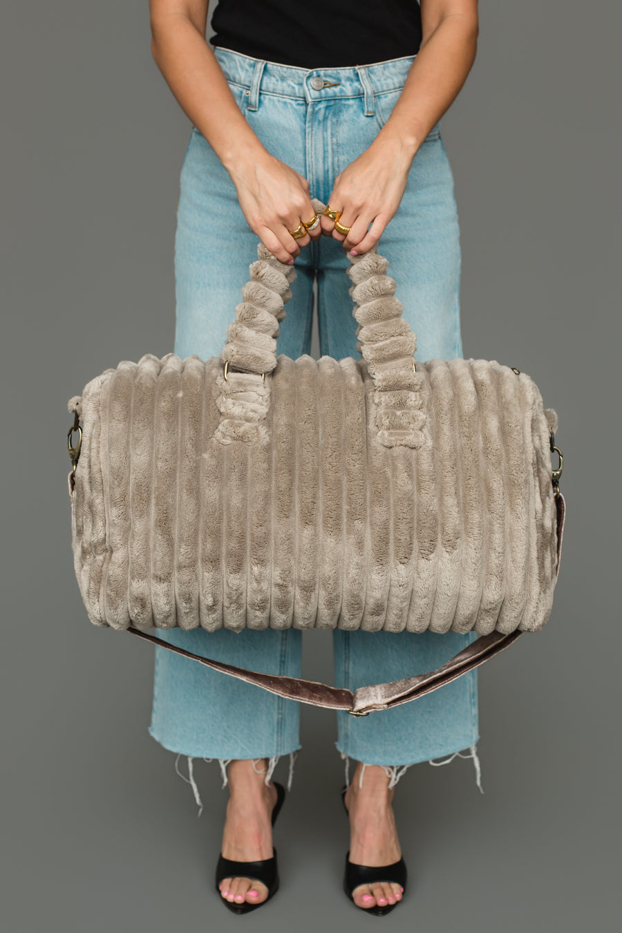 A person in frayed light blue jeans and black heels holds the Panache Apparel Collection Gray Super Soft Ribbed Duffel, featuring short handles and a removable shoulder strap, against a plain gray background.