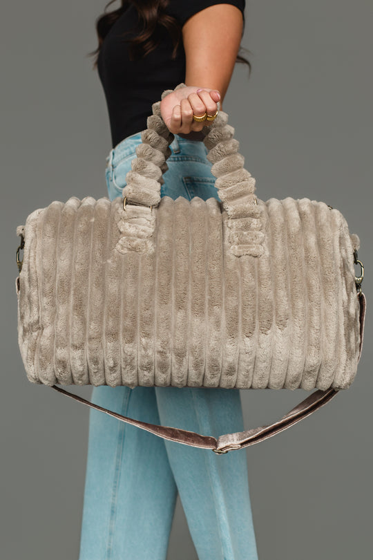 A person wearing a black shirt and blue jeans holds the Panache Apparel Collection Gray Super Soft Ribbed Duffel, featuring round handles and a shoulder strap, against a gray background.