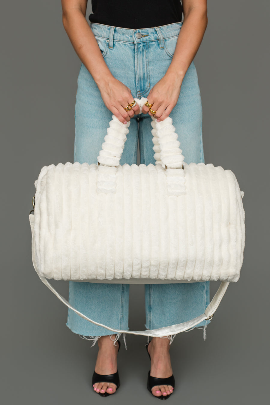 A person in jeans and black sandals holds the Panache Apparel Collection Cream Super Soft Ribbed Duffel, featuring textured handles, against a plain gray background.