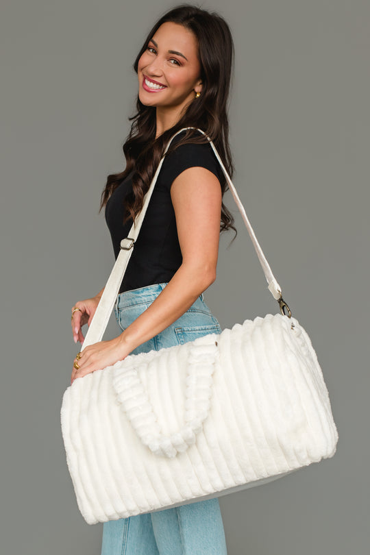 A woman wearing a black top and light blue jeans smiles while carrying the Panache Apparel Collection Cream Super Soft Ribbed Duffel, featuring both short handles and a long shoulder strap, against a plain gray background.