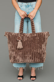 A person in light blue jeans and black sandals holds the Panache Apparel Collection Brown Super Soft Ribbed Tote, featuring thick handles, a tassel accent, and plush ribbed fabric, against a plain gray background.