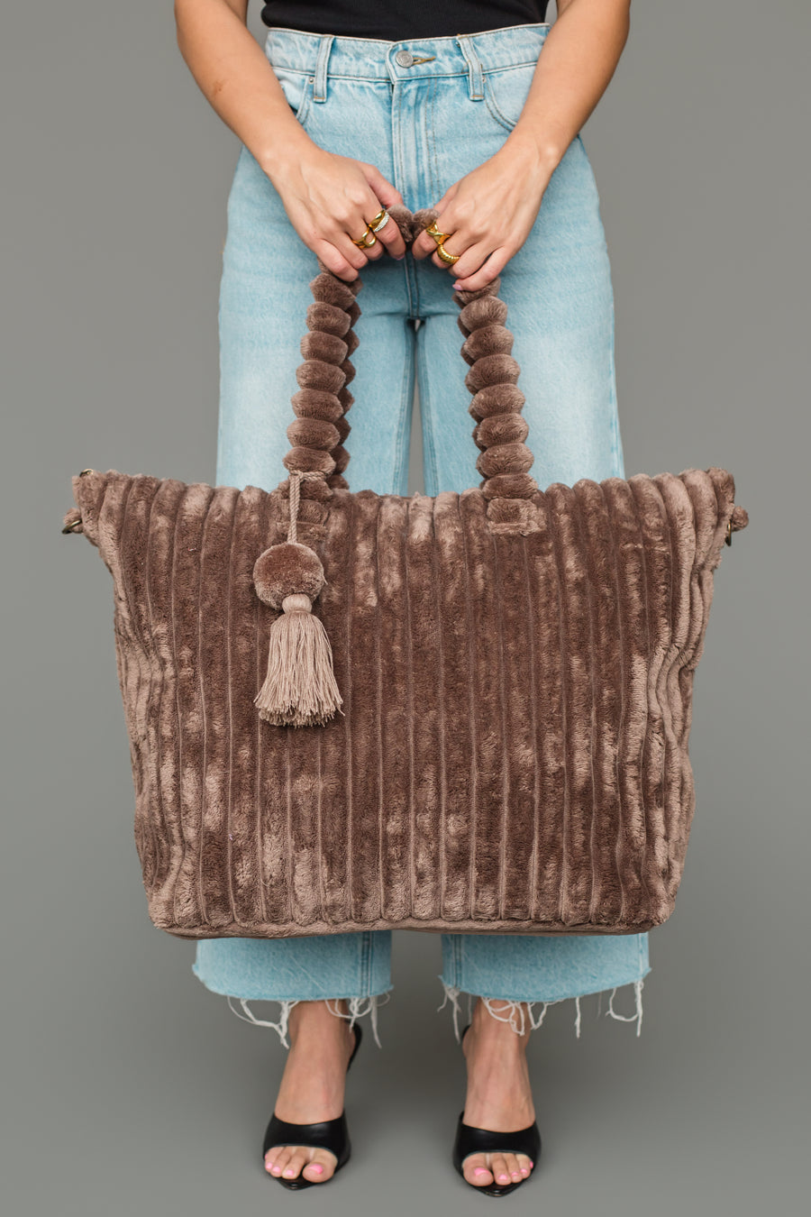 A person in light blue jeans and black sandals holds the Panache Apparel Collection Brown Super Soft Ribbed Tote, featuring thick handles, a tassel accent, and plush ribbed fabric, against a plain gray background.