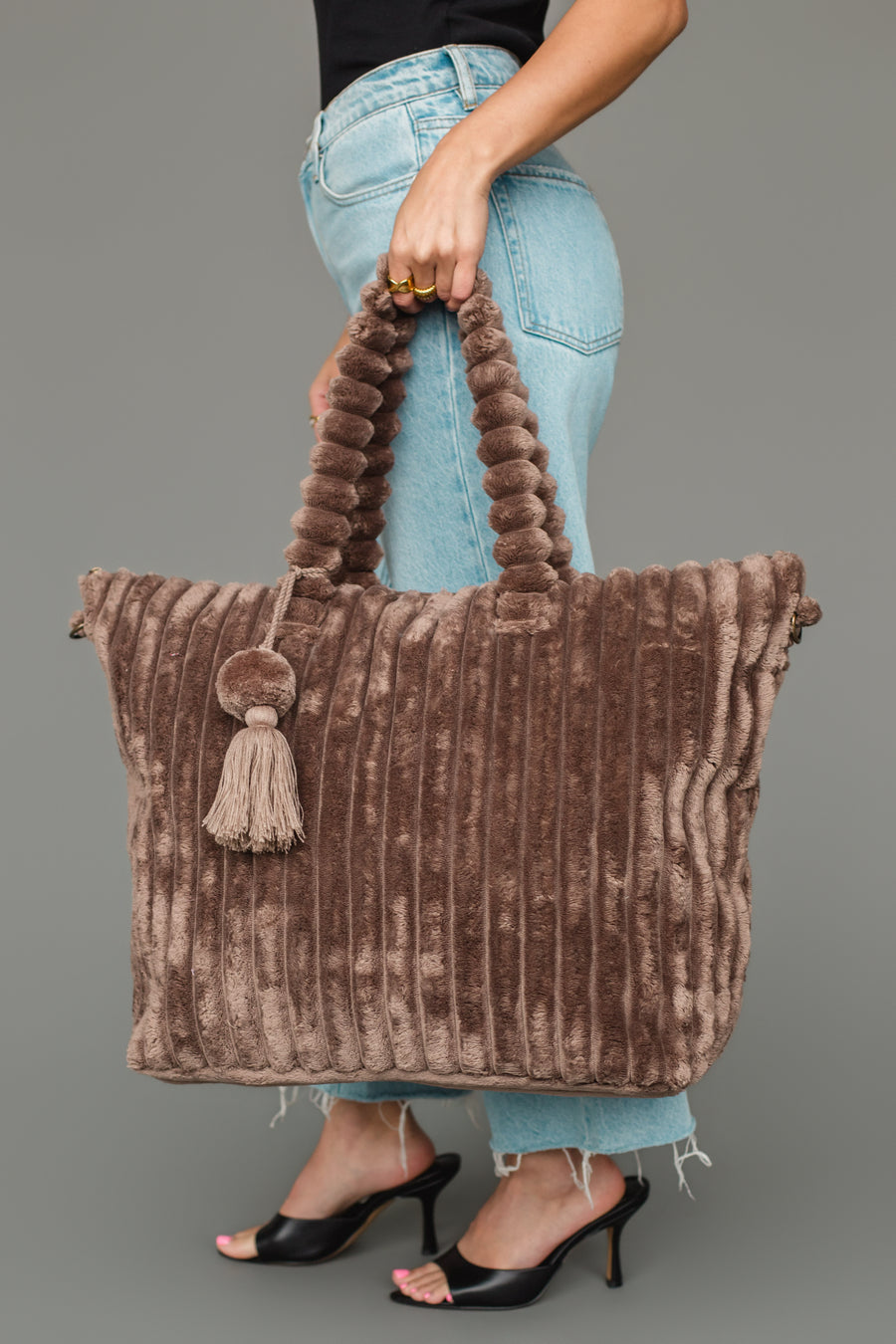 A person is holding the Panache Apparel Collection’s Brown Super Soft Ribbed Tote, featuring a plush, ridged design and a tassel. They are dressed in blue jeans, a black top, and black high-heeled sandals.