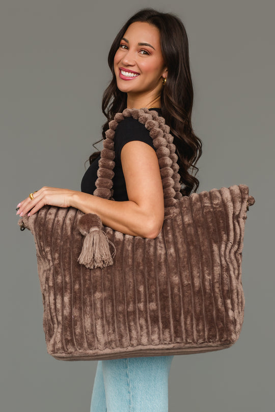 A woman smiles as she holds the Panache Apparel Collection Brown Super Soft Ribbed Tote, featuring textured handles and a tassel detail, while wearing a black shirt and light jeans against a plain gray background.