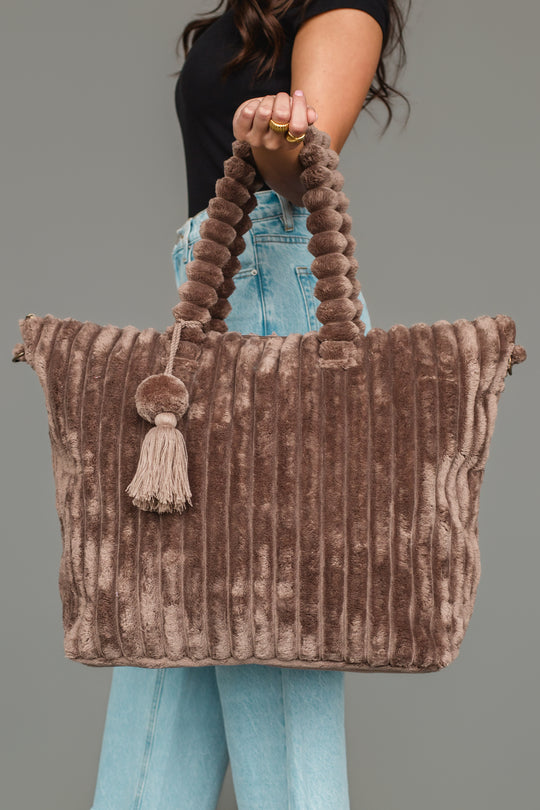 A woman in a black top and light blue jeans holds the Panache Apparel Collection’s Brown Super Soft Ribbed Tote, featuring spiral handles and a tasseled pom-pom accessory. The bag looks soft and plush.