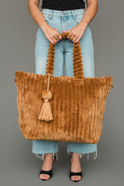 A person in jeans and black sandals holds the Panache Apparel Collection Mustard Super Soft Ribbed Tote, featuring a twisted handle and tassel detail, against a gray background.