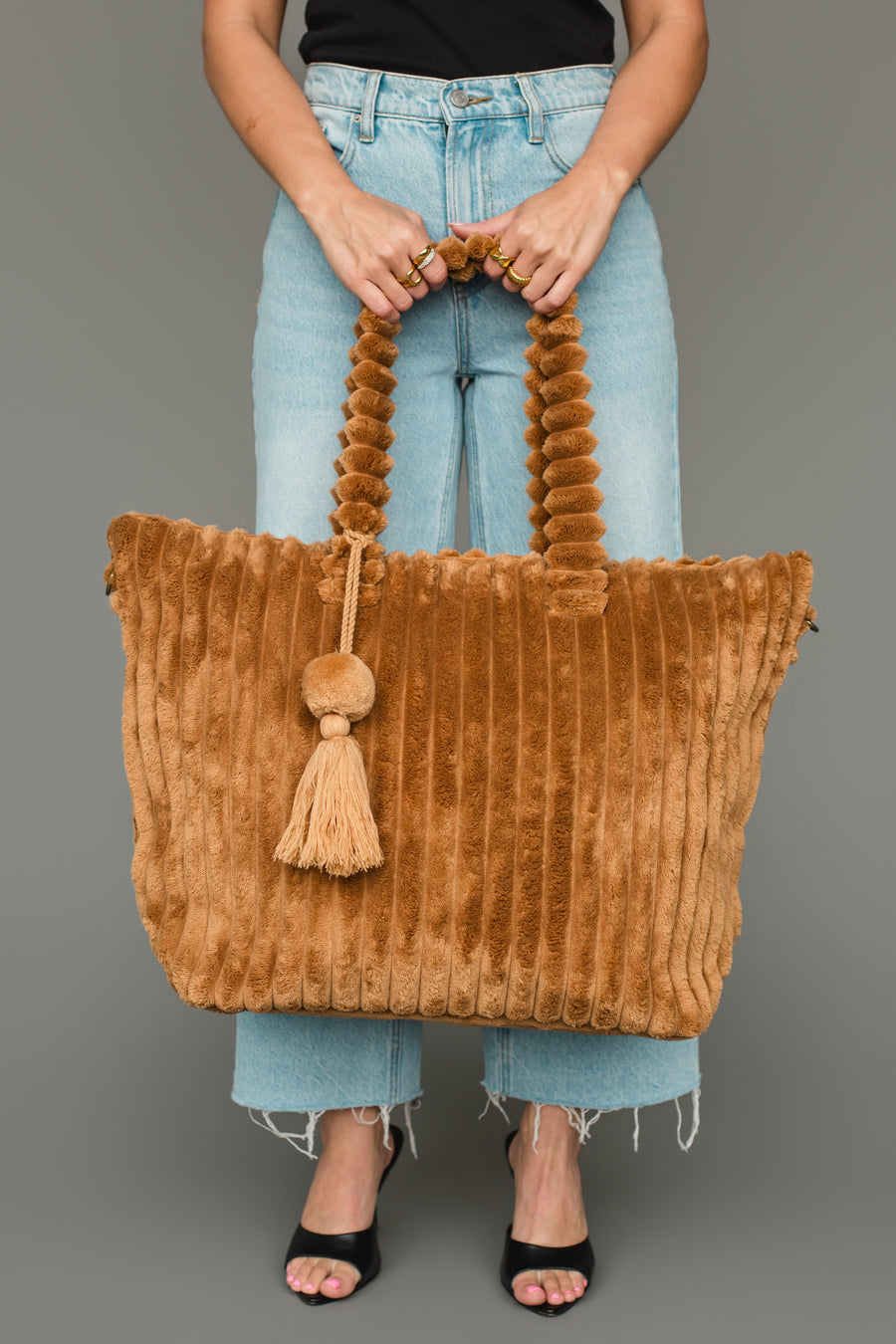 A person in jeans and black sandals holds the Panache Apparel Collection Mustard Super Soft Ribbed Tote, featuring a twisted handle and tassel detail, against a gray background.