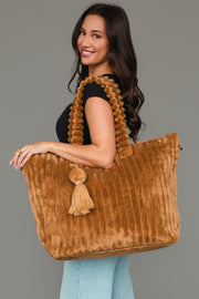 A woman with long brown hair smiles in a black top and blue jeans, holding the mustard Super Soft Ribbed Tote from Panache Apparel Collection, which features textured handles and a tassel, against a plain gray background.