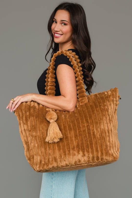A woman with long brown hair smiles in a black top and blue jeans, holding the mustard Super Soft Ribbed Tote from Panache Apparel Collection, which features textured handles and a tassel, against a plain gray background.