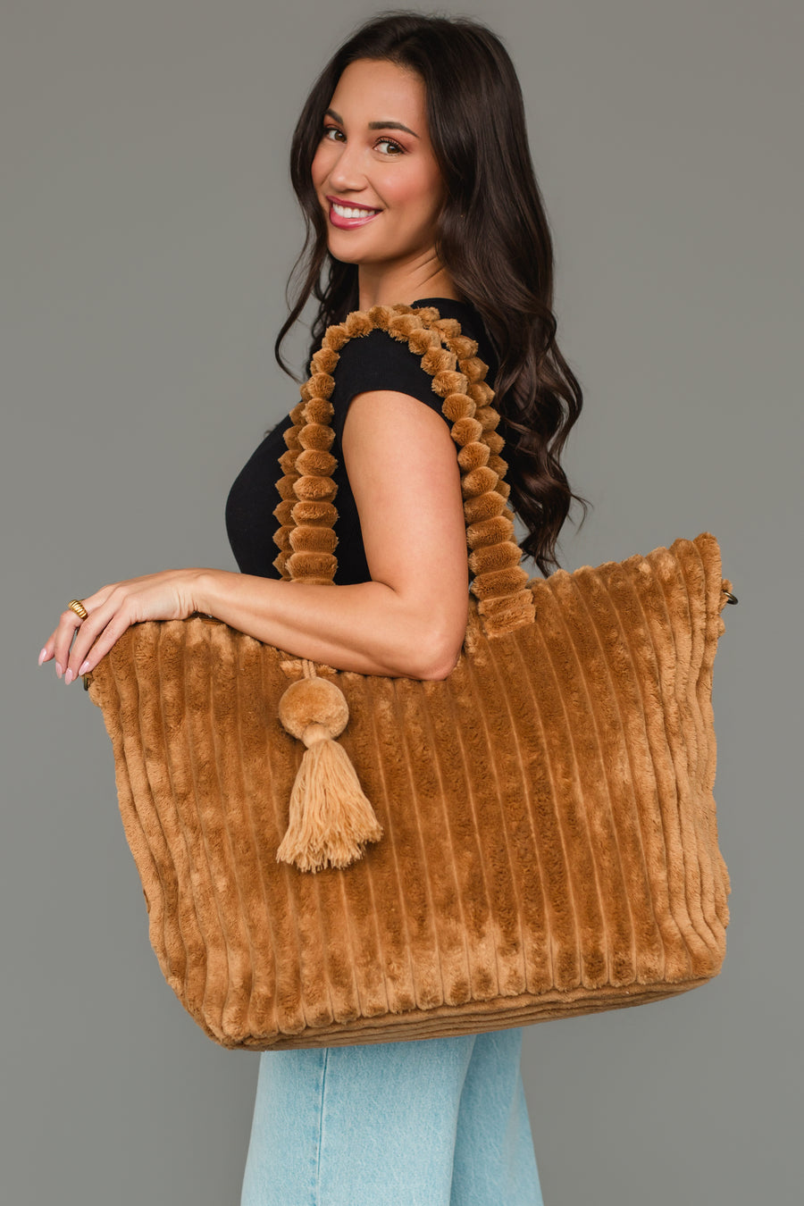 A woman with long brown hair smiles in a black top and blue jeans, holding the mustard Super Soft Ribbed Tote from Panache Apparel Collection, which features textured handles and a tassel, against a plain gray background.
