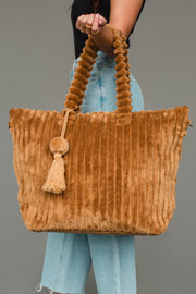 A person wearing light blue jeans holds the Panache Apparel Collection Mustard Super Soft Ribbed Tote, featuring plush vertical stripes, a round handle, and a tassel accent, against a plain gray background.