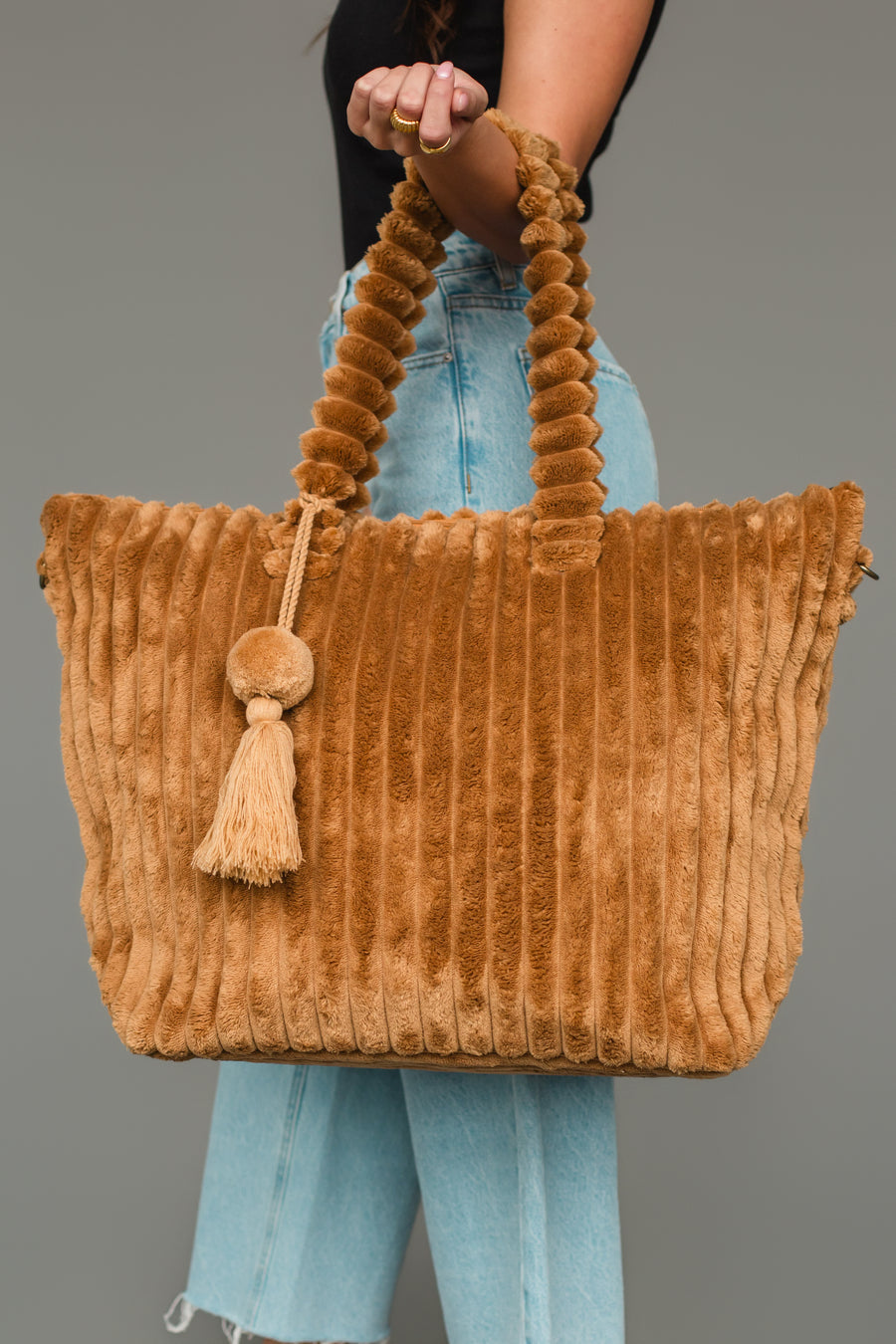 A person wearing light blue jeans holds the Panache Apparel Collection Mustard Super Soft Ribbed Tote, featuring plush vertical stripes, a round handle, and a tassel accent, against a plain gray background.