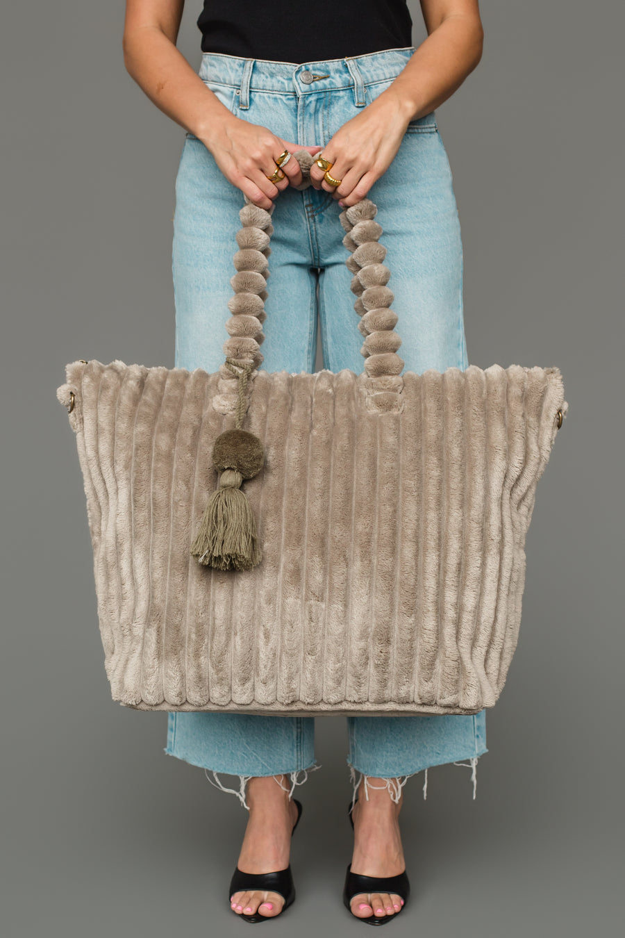 A person in jeans and a black top holds the Panache Apparel Collection Gray Super Soft Ribbed Tote, featuring thick handles and tassel accents, while standing against a plain gray background.