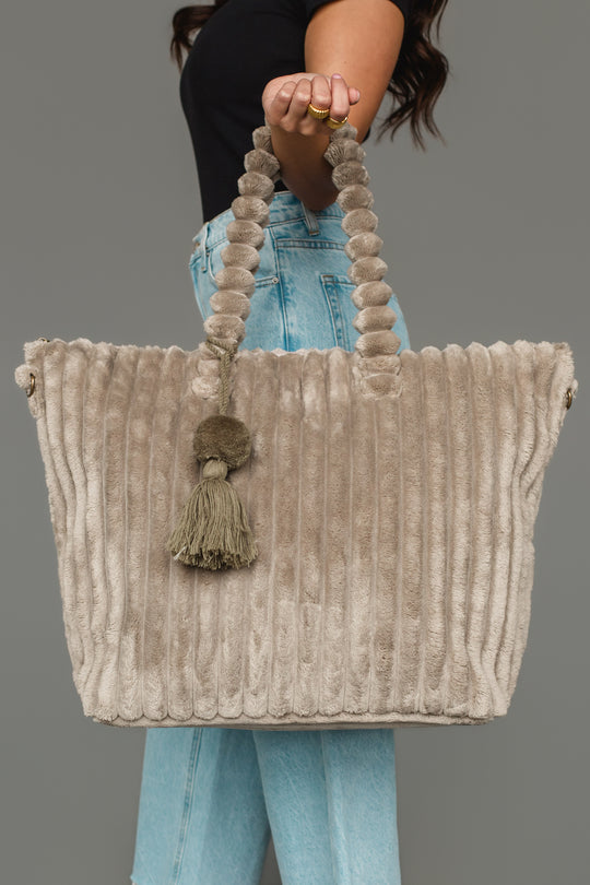 A woman in a black top and blue jeans holds the Panache Apparel Collection Gray Super Soft Ribbed Tote, featuring plush textured handles and a decorative tassel, against a plain gray background.