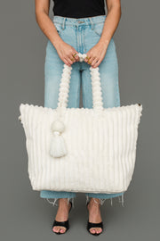A woman wearing a black top, light blue jeans, and black heels holds the Panache Apparel Collection Cream Super Soft Ribbed Tote, featuring beaded handles and a tassel, in front of a gray background.