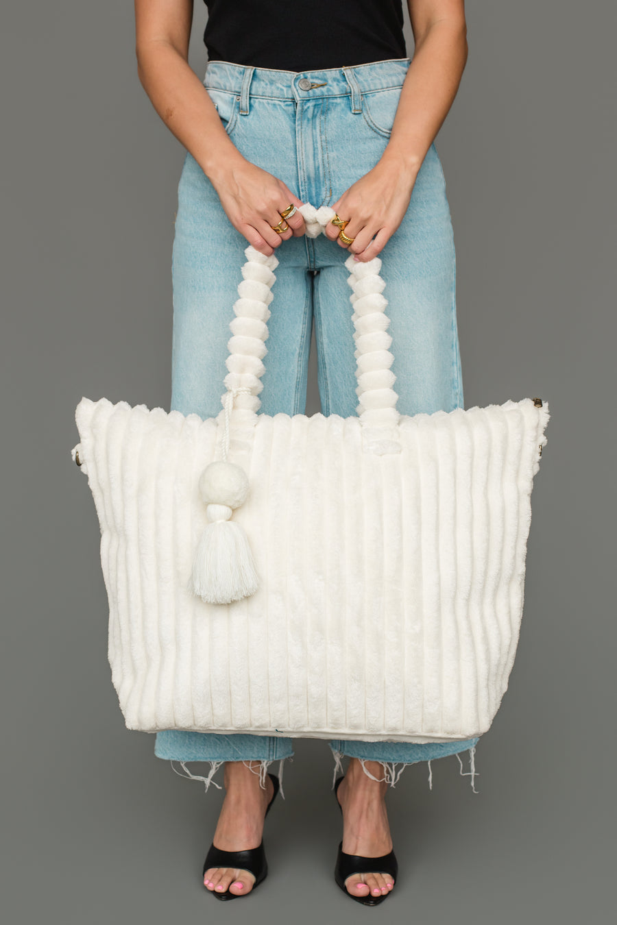 A woman wearing a black top, light blue jeans, and black heels holds the Panache Apparel Collection Cream Super Soft Ribbed Tote, featuring beaded handles and a tassel, in front of a gray background.