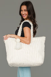 A woman in a black top and light jeans smiles while holding the Cream Super Soft Ribbed Tote by Panache Apparel Collection, featuring textured fabric and pom-pom details, over her shoulder against a plain gray background.