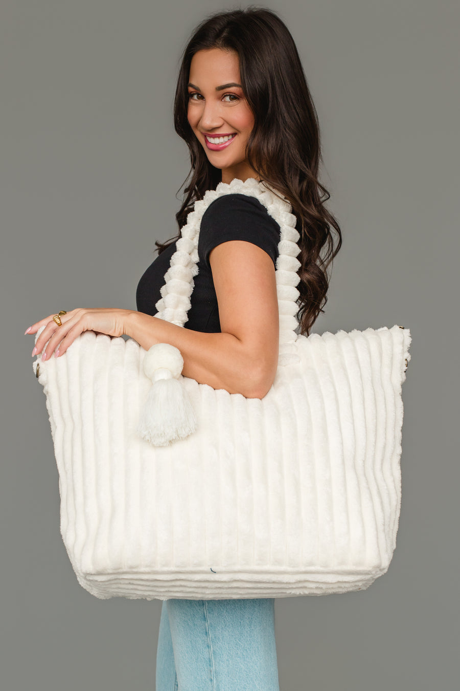 A woman in a black top and light jeans smiles while holding the Cream Super Soft Ribbed Tote by Panache Apparel Collection, featuring textured fabric and pom-pom details, over her shoulder against a plain gray background.