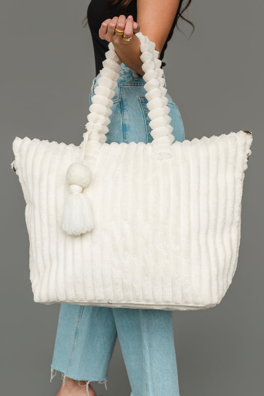 A woman wearing a black top and blue jeans holds the Panache Apparel Collection Cream Super Soft Ribbed Tote, featuring twisted handles and decorative tassels, against a plain gray background.
