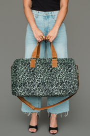 A person wearing light blue jeans and black sandals holds the Panache Apparel Collection Green & Black Leopard Duffel, featuring brown handles and a shoulder strap, against a gray background.