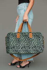 A person in blue jeans and black heels holds the Panache Apparel Collection Green & Black Leopard Duffel against a gray background.