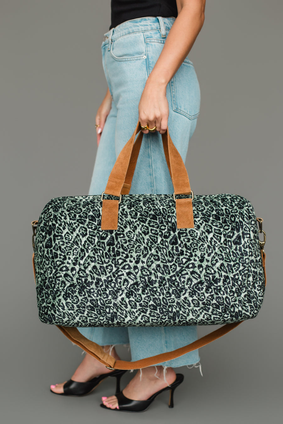 A person in blue jeans and black heels holds the Panache Apparel Collection Green & Black Leopard Duffel against a gray background.