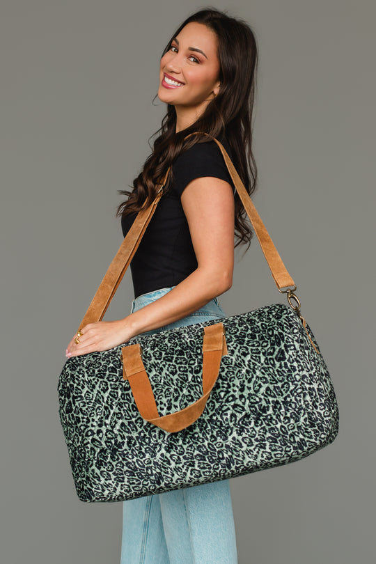 A woman in a black top and light jeans smiles while holding the Panache Apparel Collection Green & Black Leopard Duffel with tan straps over her shoulder, standing against a plain gray background.