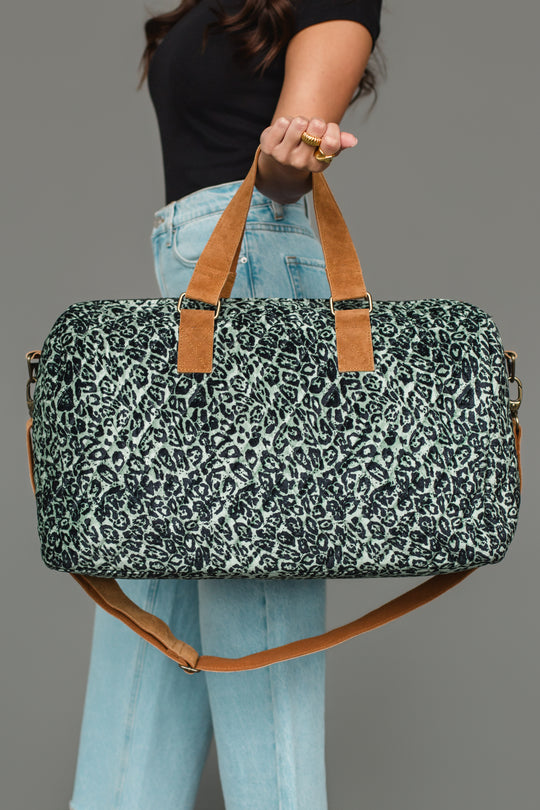 A person in a black top and light blue jeans holds the Panache Apparel Collection Green & Black Leopard Duffel, featuring brown straps and a shoulder strap, against a plain gray background.