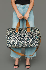 A person is holding the Black, Cream & Blue Leopard Duffel from Panache Apparel Collection, featuring brown straps. They are dressed in light blue jeans, a black top, and black open-toe heels, standing against a plain gray background.