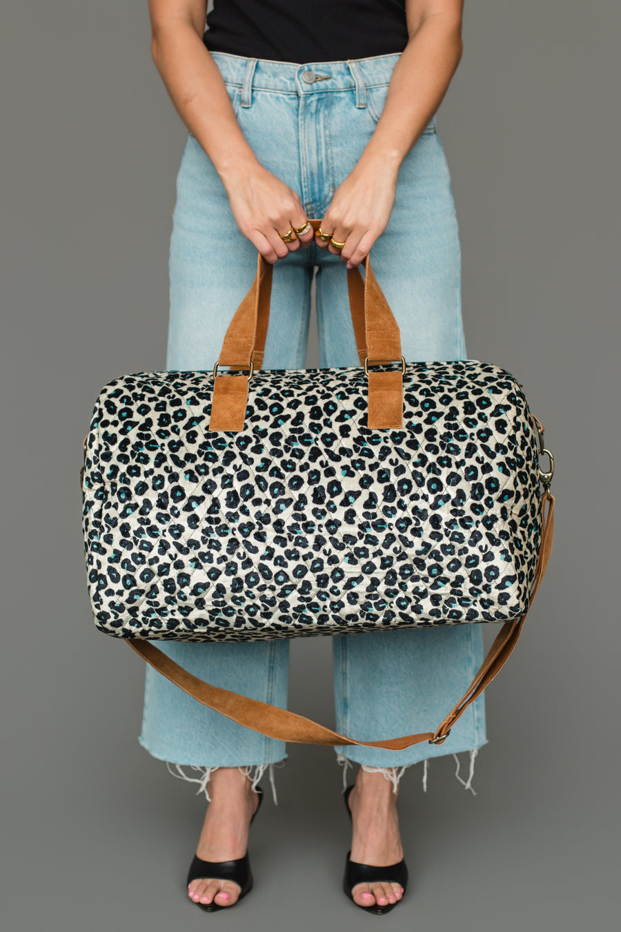 A person is holding the Black, Cream & Blue Leopard Duffel from Panache Apparel Collection, featuring brown straps. They are dressed in light blue jeans, a black top, and black open-toe heels, standing against a plain gray background.