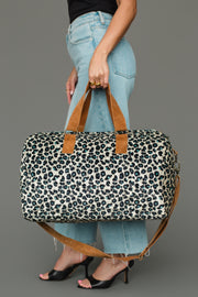 A person in blue jeans and black heels holds the Panache Apparel Collection Black, Cream & Blue Leopard Duffel, featuring brown handles and a shoulder strap, against a neutral gray background.