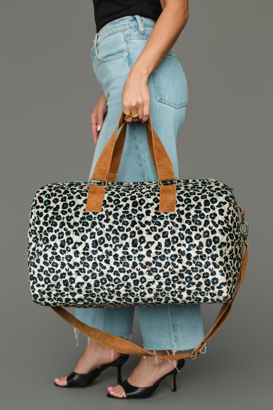 A person in blue jeans and black heels holds the Panache Apparel Collection Black, Cream & Blue Leopard Duffel, featuring brown handles and a shoulder strap, against a neutral gray background.
