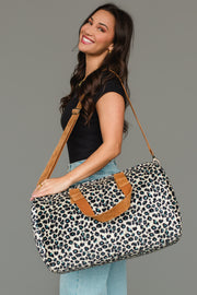 A smiling woman in a black top and light blue jeans carries the Panache Apparel Collection Black, Cream & Blue Leopard Duffel over her shoulder, featuring tan straps, while standing against a plain grey background.