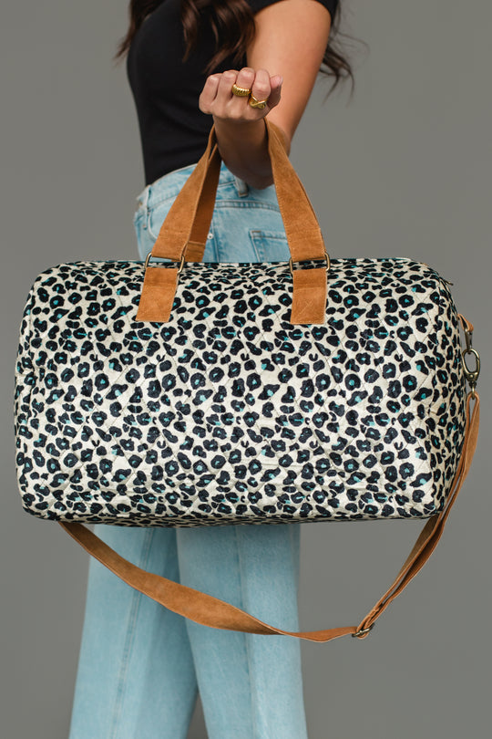 A woman wearing a black top and light jeans holds the Panache Apparel Collection Black, Cream & Blue Leopard Duffel, featuring tan handles and a matching tan shoulder strap, against a plain gray background.