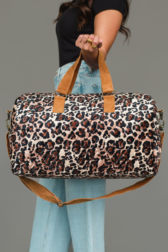 A person wearing a black top and light blue jeans holds the Panache Apparel Collection Brown & Cream Leopard Duffel, featuring tan handles and a shoulder strap, against a plain gray background.