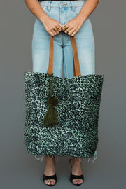 Wearing light blue jeans and black heels, a person holds the Panache Apparel Collection Green & Black Leopard Tote, featuring brown handles and a green tassel, against a plain gray background.