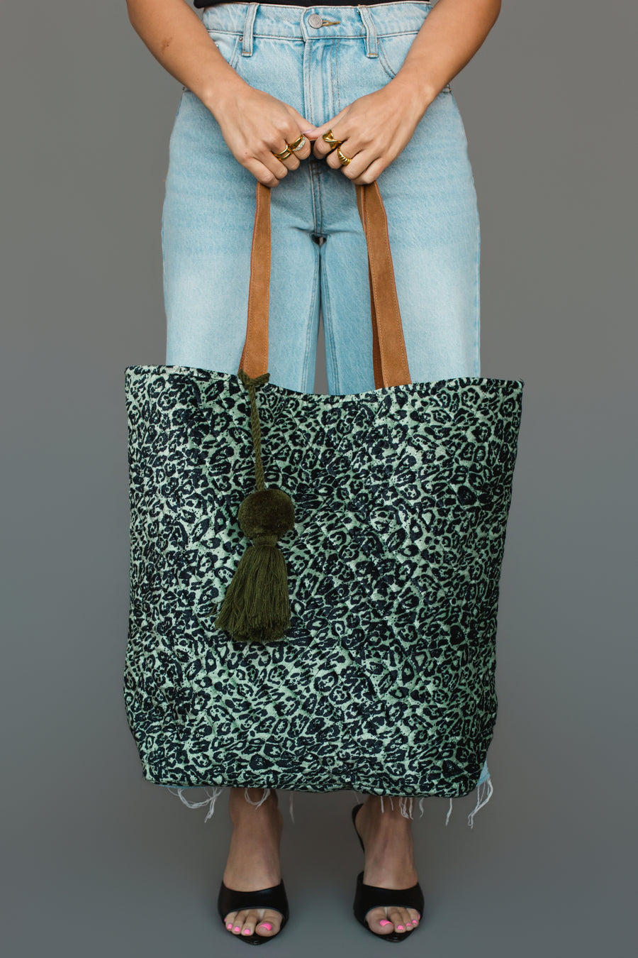 Wearing light blue jeans and black heels, a person holds the Panache Apparel Collection Green & Black Leopard Tote, featuring brown handles and a green tassel, against a plain gray background.