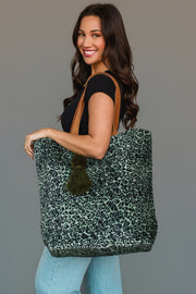 A woman smiles while holding the Panache Apparel Collection Green & Black Leopard Tote, featuring brown straps and a dark green tassel, paired with a black top and light blue jeans.