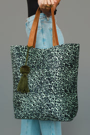 A person holds the Green & Black Leopard Tote from Panache Apparel Collection, featuring brown handles and a green tassel, while wearing light blue jeans and a black top.