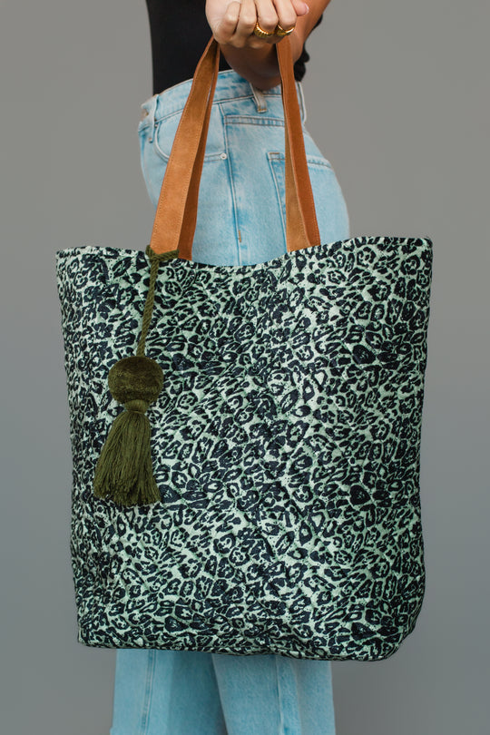 A person holds the Green & Black Leopard Tote from Panache Apparel Collection, featuring brown handles and a green tassel, while wearing light blue jeans and a black top.