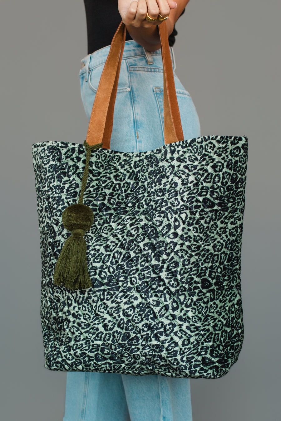 A person holds the Green & Black Leopard Tote from Panache Apparel Collection, featuring brown handles and a green tassel, while wearing light blue jeans and a black top.